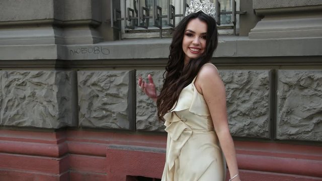 beautiful girl, brunette, in a light long dress with a deep neckline, long hair and bright makeup, crown on her head, posing and smiling in front of the camera. takes place on the street near building