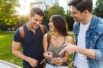 Cheery happy young friends walking in nature green park outdoors using mobile phone.