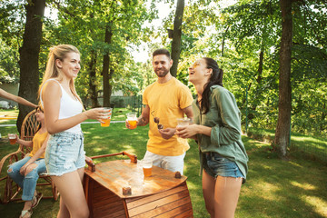 Group of happy friends having beer and barbecue party at sunny day. Resting together outdoor in a forest glade or backyard. Celebrating and relaxing, laughting. Summer lifestyle, friendship concept.