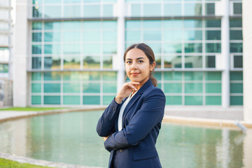 Satisfied successful business leader posing outside. Beautiful Latin woman wearing formal suit, standing near office building, touching chin and looking at camera. Young business woman concept
