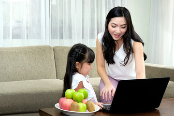 Asian young mother sitting on sofa in the living room with her daughter while is teaching the use of laptop. Relaxing days in a warm family.