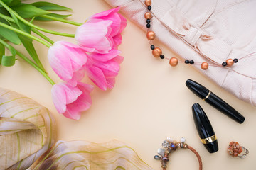 A composition of five buds of pink flowers, fragments of a dress and scarf, cosmetics, necklaces, rings and bracelets. Top view on a beige background