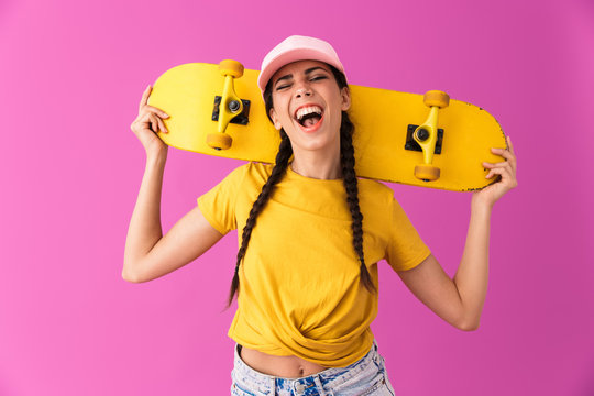 Image of young teenage woman wearing cap laughing and holding skateboard on her shoulders