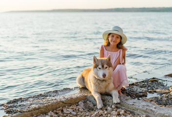 Little beautiful girl in pink dress and hat are playing with siberian husky dog near the sea. young girl are walking on the coast, at the sunset time. © Tetiana