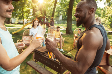 Group of happy friends having beer and barbecue party at sunny day. Resting together outdoor in a forest glade or backyard. Celebrating and relaxing, laughting. Summer lifestyle, friendship concept.