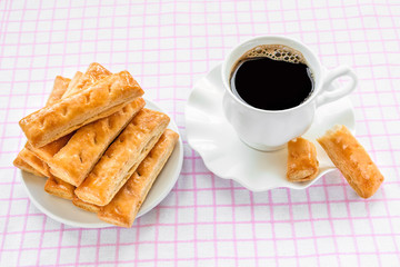Cup with hot black coffee and sweet puff pastry cookies on a white saucer over white pink checkered tablecloth. Tasty breakfast or snack.