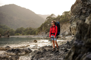 Woman standing on the rocks on the coastline on the sea with hiking backpack