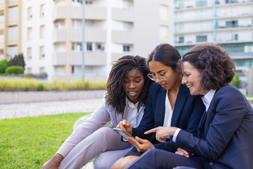 Fototapeta premium Excited businesswoman watching content on phone and showing screen to female colleagues. Women wearing office suits, sitting on grass during work break. Business communication concept