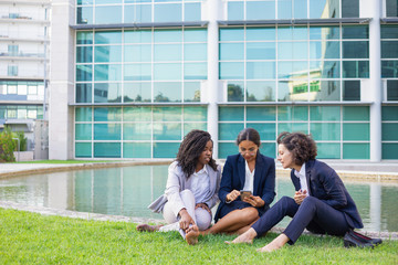 Barefoot multiethnic businesswomen with smartphone outdoor. Young female colleagues sitting on green lawn and using mobile phone. Technology concept