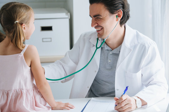 Male Doctor Examining A Child Girl, That Sits On A Table In A Hospital