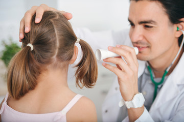 Male doctor examining little girl's ear at hospital, using flashlight