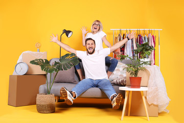 Young married couple moving in new apartment, sitting between many boxes and things. New home concept. Bearded man and blonde woman resting on couch over yellow wall. Background, copy space, close up.