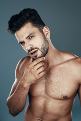 Obraz premium Handsome young shirtless man looking at camera and keeping hand on chin while standing against grey background