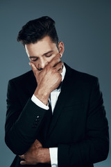Tired young man in bow tie covering mouth with hand while standing against grey background