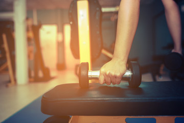 Hands are holding a black dumbbell in the gym with a backdrop of fitness room and sun light.