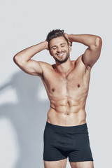 Handsome young shirtless man smiling and keeping hands behind head while standing against grey background