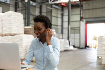 Female manager talking on headset while using laptop at desk in warehouse