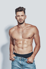 Handsome young shirtless man looking at camera and taking off his jeans while standing against grey background