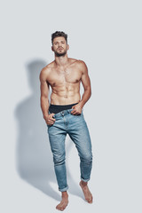 Full length of handsome young shirtless man looking at camera while standing against grey background