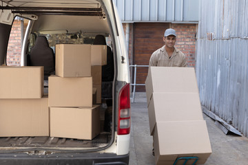 Delivery man carrying cardboard boxes on trolley near van outside the warehouse