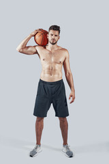 Fototapeta na wymiar Full length of handsome young man in sports clothing carrying basketball ball and looking at camera while standing against grey background