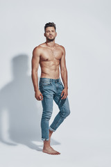 Fototapeta premium Full length of handsome young shirtless man looking at camera while standing against grey background