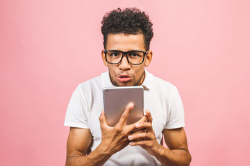 Excited african American male in glasses isolated on pink studio background hold tablet get good message, surprised black man receive get to know about sale offer or discount on pad.