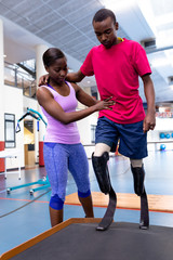 Female physiotherapist helping disabled man walk with prosthetic leg on ramp in sports center
