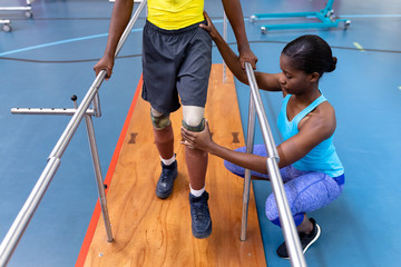 Fototapeta na wymiar Physiotherapist assisting disabled man walk with parallel bars in sports center
