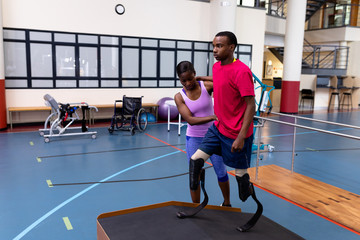 Physiotherapist helping disabled man walk with prosthetic leg on ramp in sports center