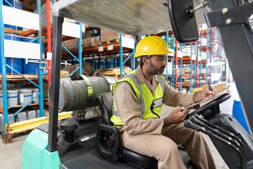 Male staff writing on clipboard while sitting on forklift in warehouse