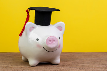 Smiling pink piggy bank wearing graduated hat on wooden table, yellow background and copy space, education fund, Scholarships, university cost and expense or saving for student loan concept