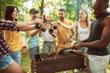 Group of happy friends having beer and barbecue party at sunny day. Resting together outdoor in a forest glade or backyard. Celebrating and relaxing, laughting. Summer lifestyle, friendship concept.