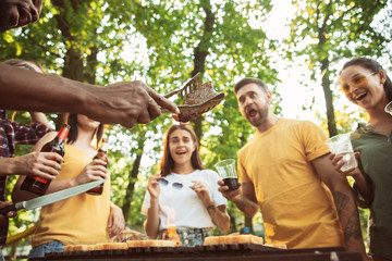 Group of happy friends having beer and barbecue party at sunny day. Resting together outdoor in a forest glade or backyard. Celebrating and relaxing, laughting. Summer lifestyle, friendship concept.