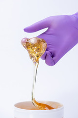 Woman's hand in a glove holds yellow sugar paste or wax for depilation close up on a white...