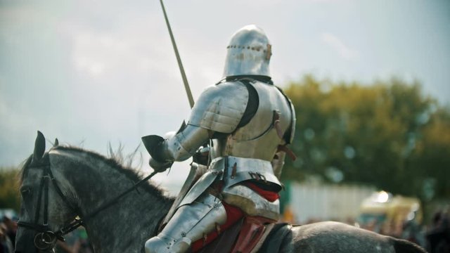 A man knight riding a horse around the battlefield and greeting the people watching behind the fence - raises his hand up in the sky holding a sword