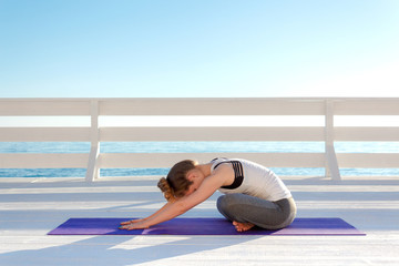 Young woman practicing yoga outdoors at white wooden seafront. Sitting in easy pose and bending...