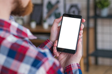 Mockup image - over shoulder view: man entrepreneur hands holding black smartphone with white blank screen in home interior. Mock up, copyspace, template, entertainment and technology concept
