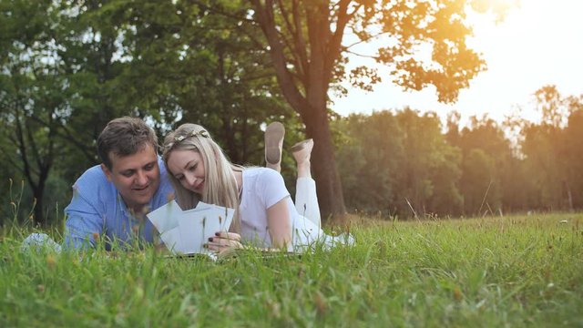 A guy and a girl are watching a family album with photos in the park on the grass.