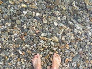 A pair of feet standing on colourful pebbels in the shallow waters of a crystal clear lake in New Zealand