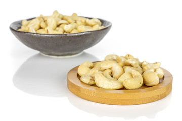 Lot of whole unsalted beige cashew on round bamboo coaster in dark ceramic bowl isolated on white background