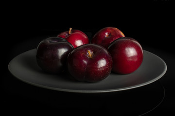 Group of five whole ripe red round plum on gray ceramic plate isolated on black glass