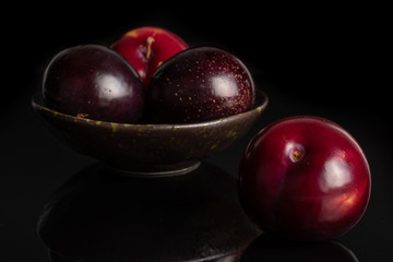 Group of four whole ripe red round plum in dark ceramic bowl isolated on black glass