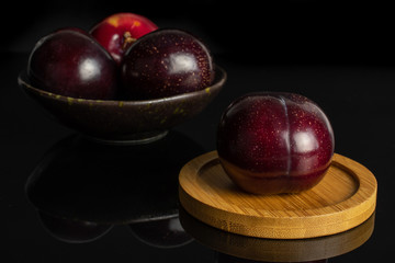 Group of four whole ripe red round plum on round bamboo coaster in dark ceramic bowl isolated on black glass