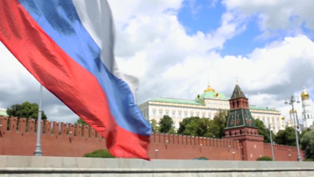 Russian flag waving from wind against backdrop of red wall with towers and green trees. Movement along Kremlin embankment in Moscow on sunny day