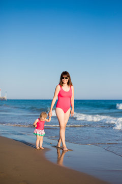 summer and travel concept - happy mother and little daughter in pink swimsuits walking at the beach