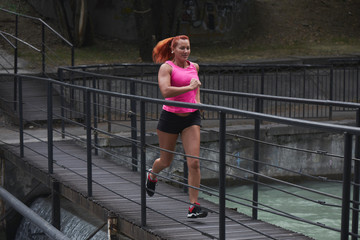 Beautiful adult red haired woman athlete in pink top and shorts makes amorning run on the city...
