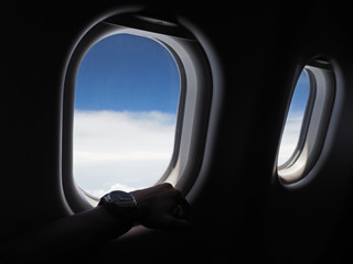 Fototapeta premium Silhouette of a male hand looking at his wrist watch with airplane window and blue sky background.