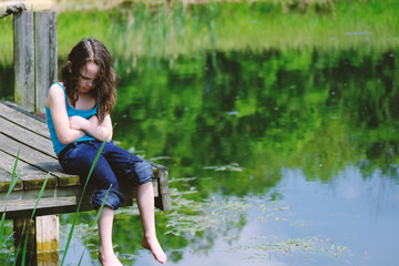 Young girl sitting on dock by rural pond water, arms crossed.  Concept of upset child.