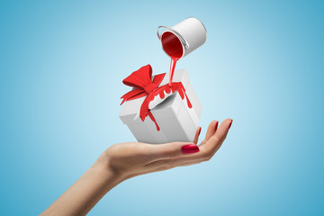 Female hand with red paint bucket turned upside down above white gift box on blue background